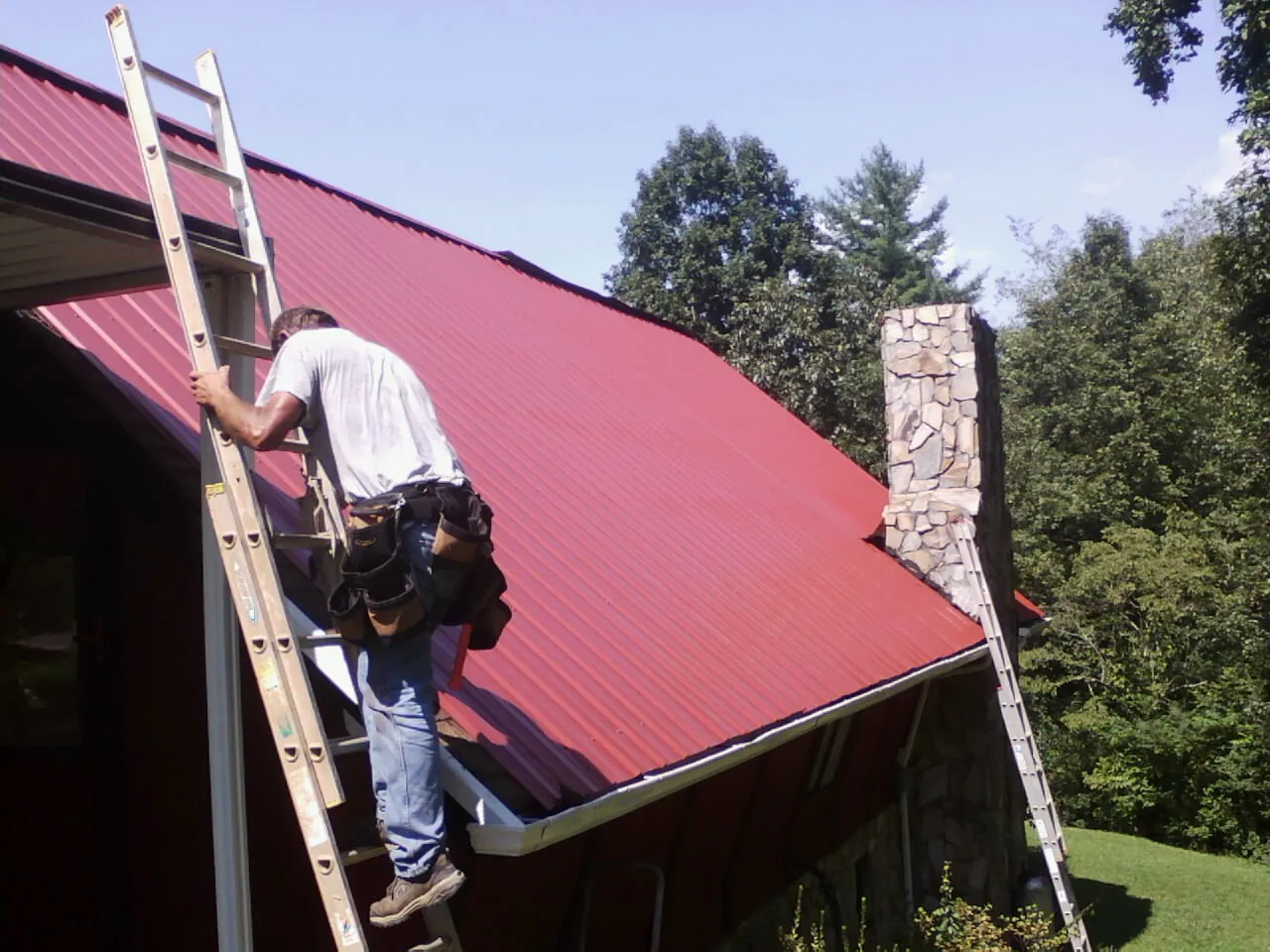 Roofer on a ladder beside a red metal roof and stone chimney — Soffit & Fascia Repair by Roofing Brothers in Elysburg